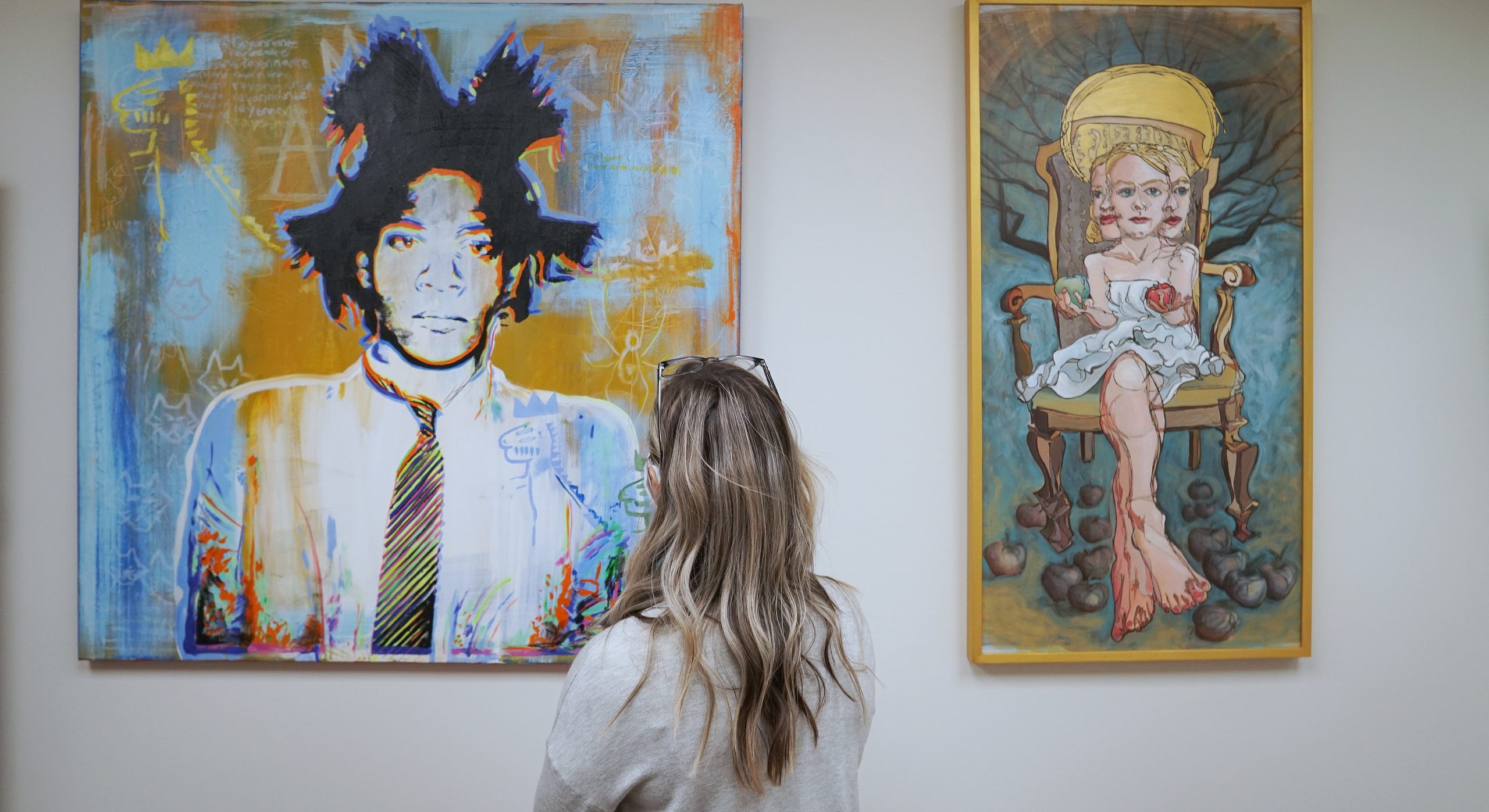 A White woman stands in front of a painting with a person with blues and yellows
