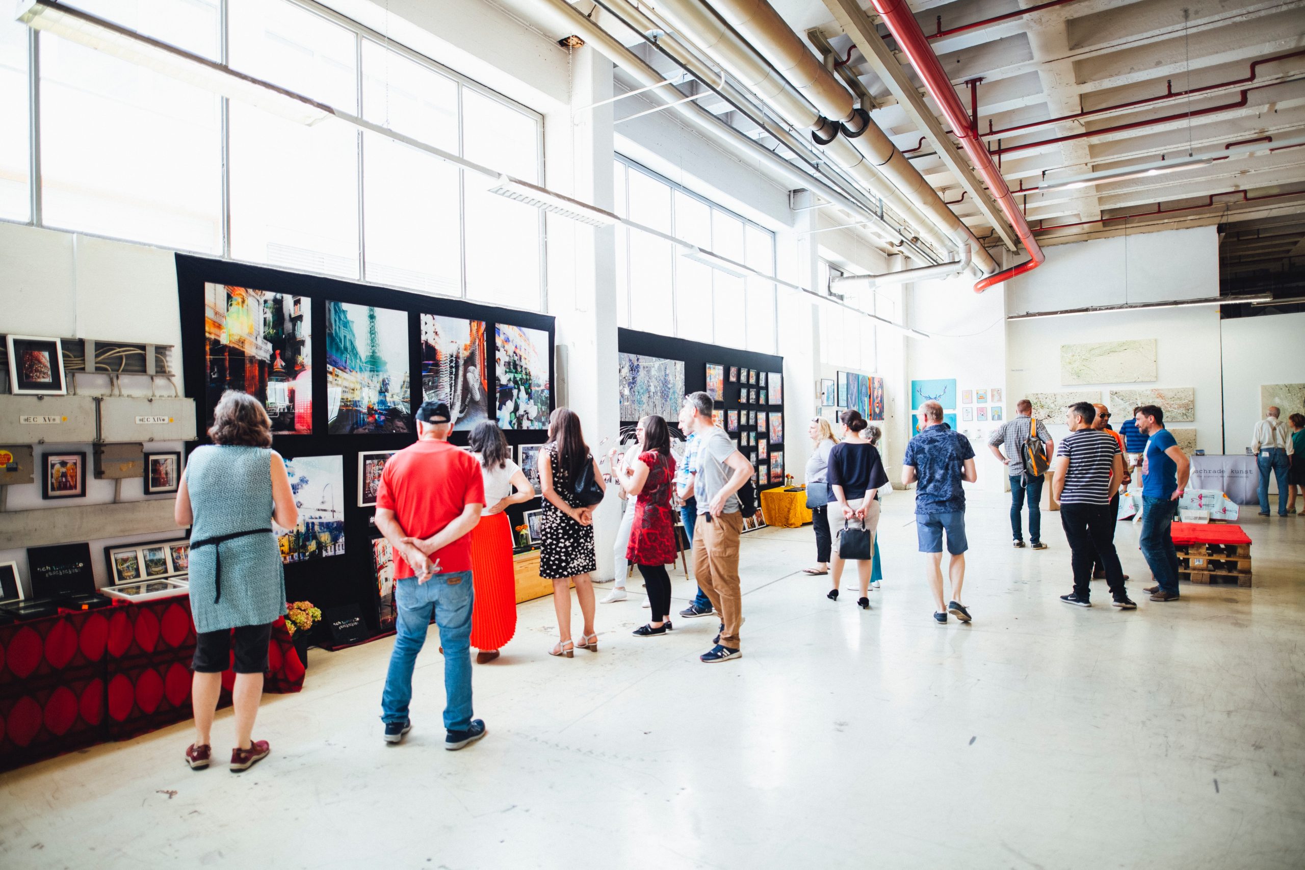 Bright open event space with several piece of art hung on walls and approximately twenty people standing in the space looking at the artwork.