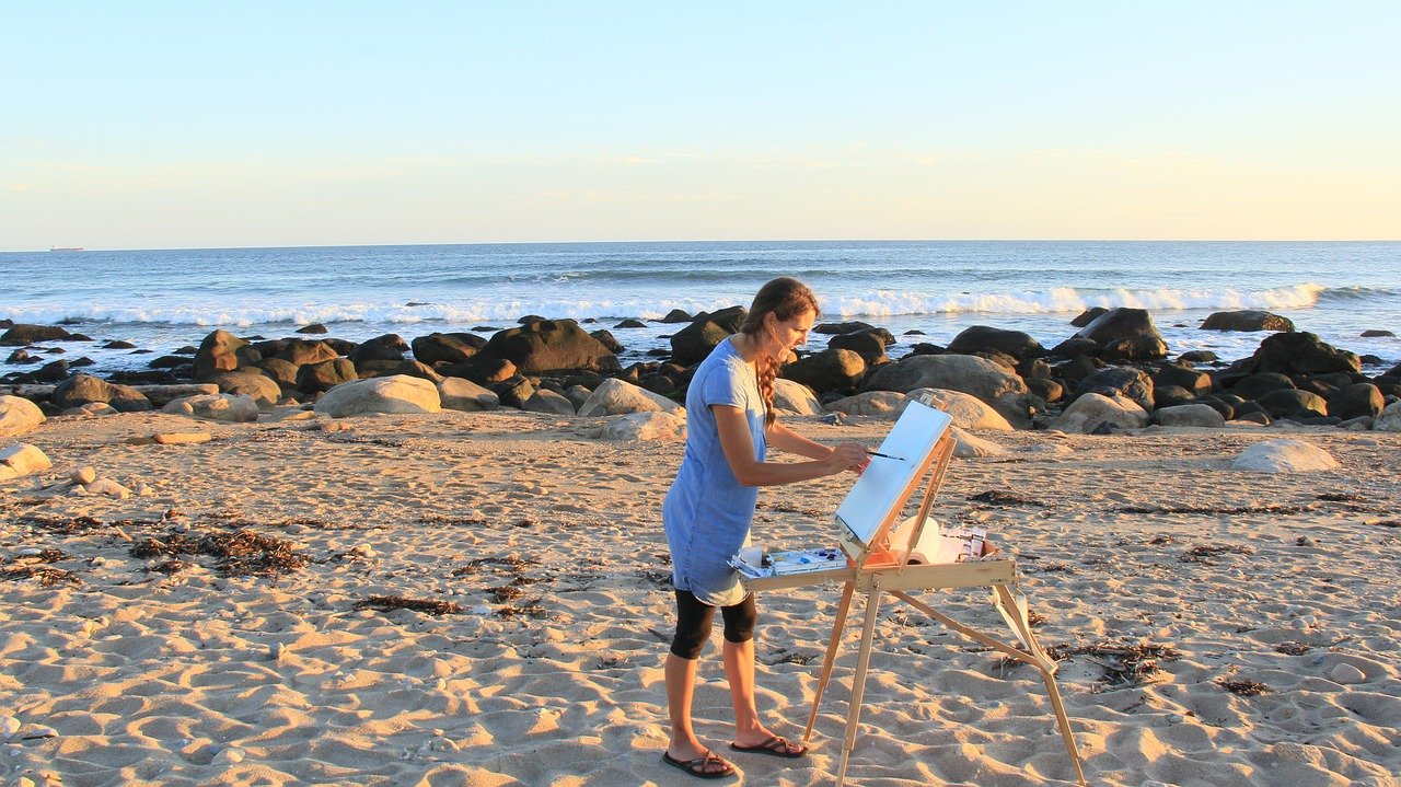 in the background there is an ocean coast line with a clear sky and a rocky beach. In the foreground there is a woman wearing gray clothes standing in front of a canvas on a stand-up easel and is painting.