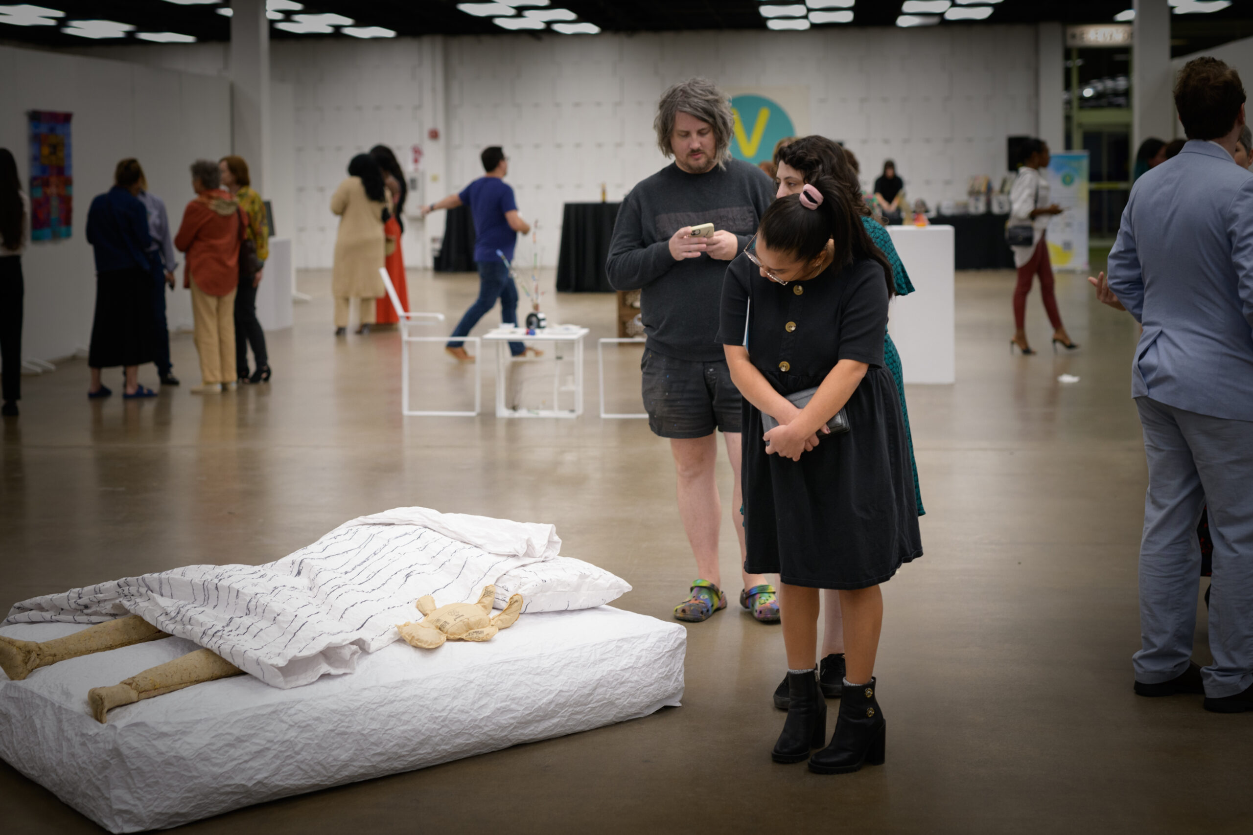 Texas Vignette a woman in a black dress and black shoes leans over to examine a sculpture of a wooden mannequin on a mattress