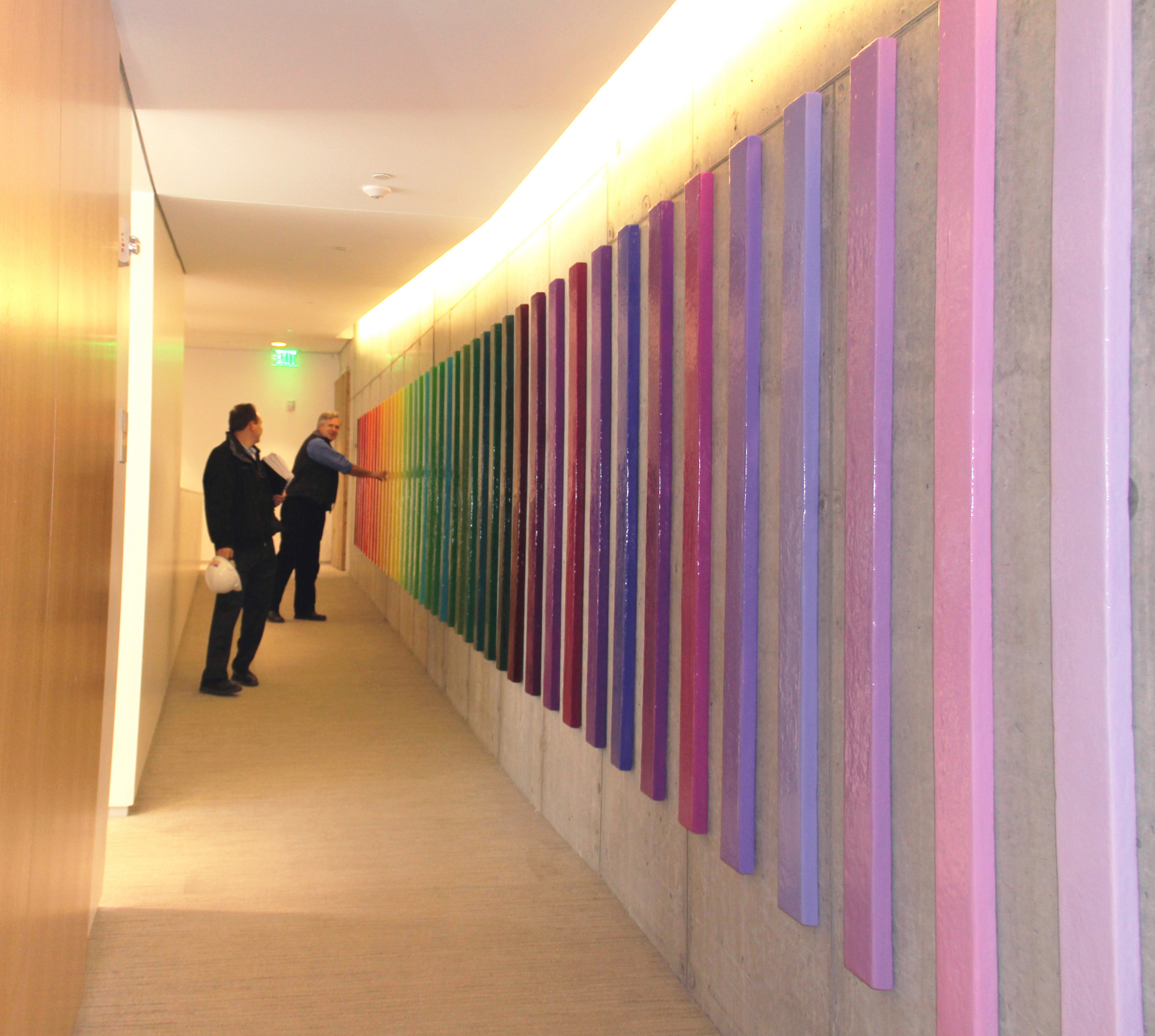 Photograph depicting a hallway with two people looking at a bluck art sculpture of multiple, colored panels hanging on the wall in rainbow order