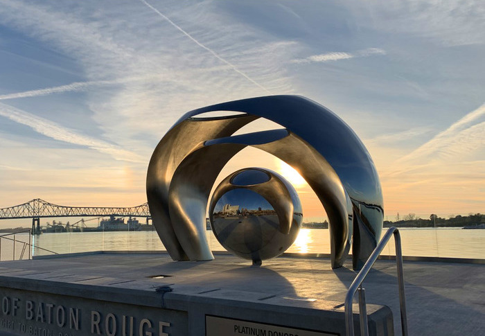 Photograph depicting a large, steel sculpture displayed outside in front of the Mississippi river during sunset