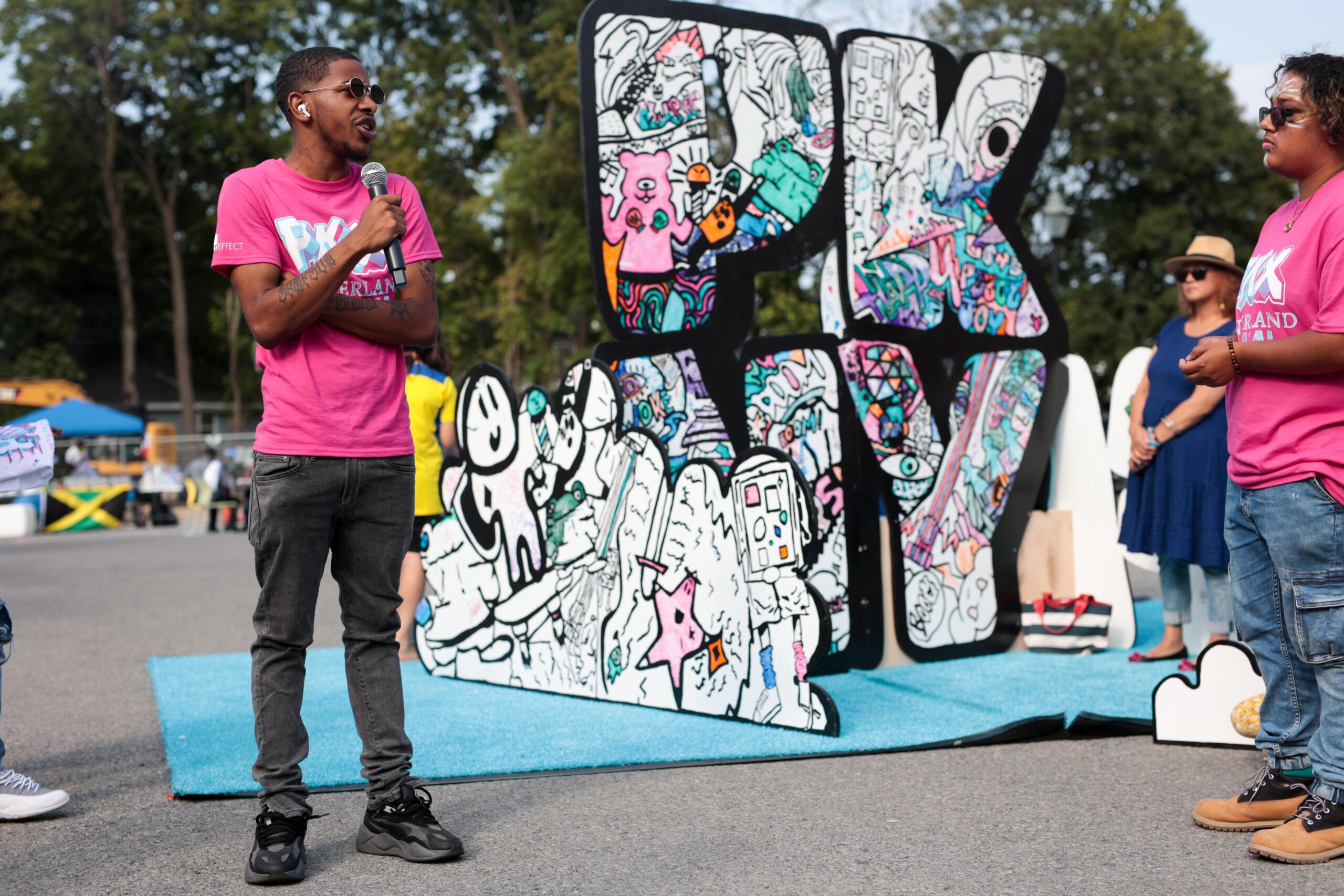 photo of a man in a pink shirt holding a microphone and talking while standing in front of an interactive art piece of the letters 