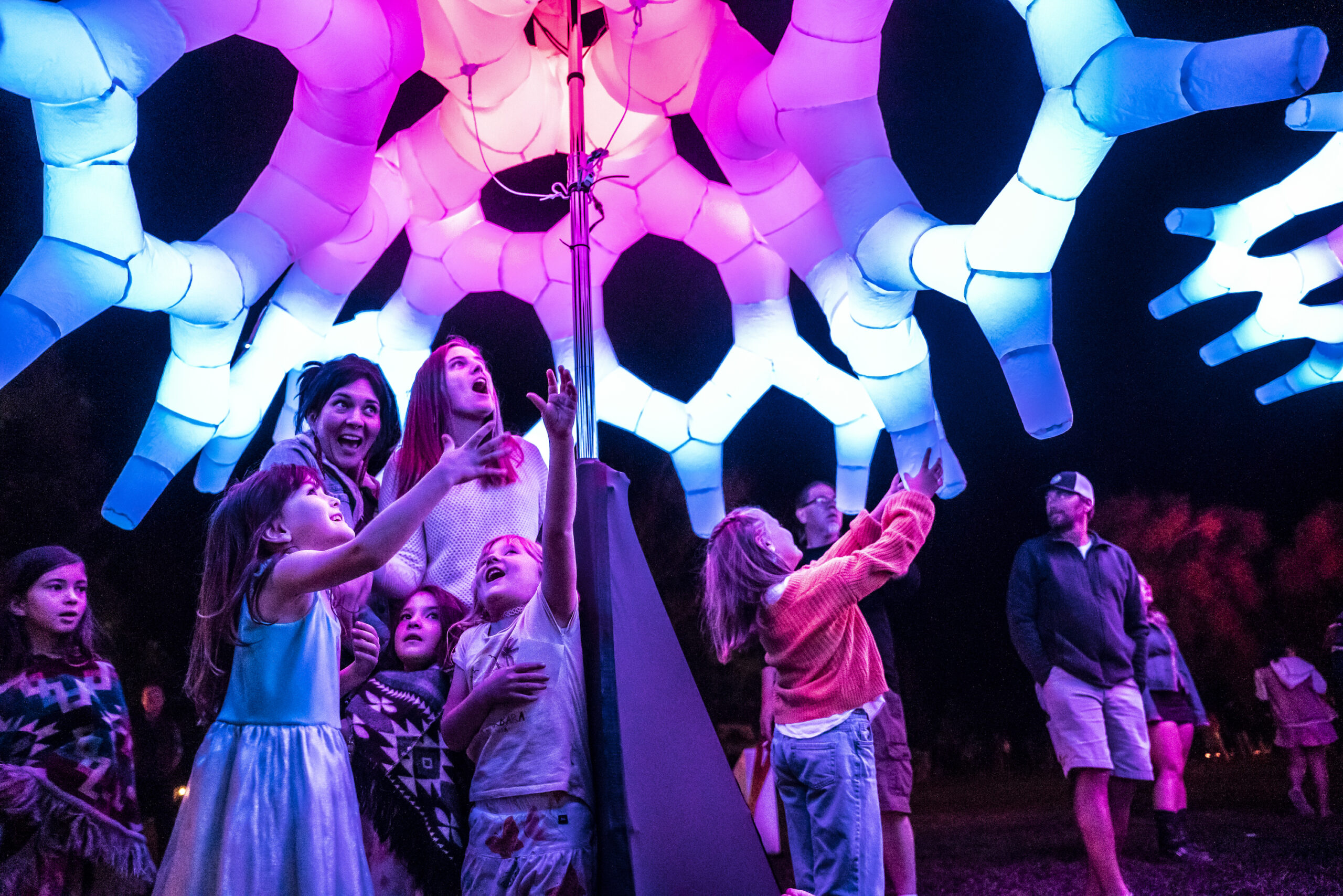 Adults and kids standing under a very colorful public art piece smiling and having fun.