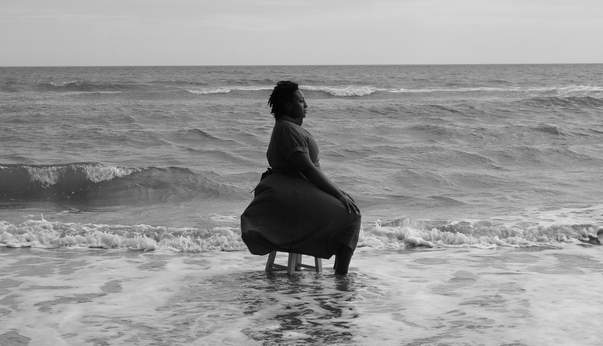 Black and white photograph of a woman sitting on a chair in the waves of a beach.