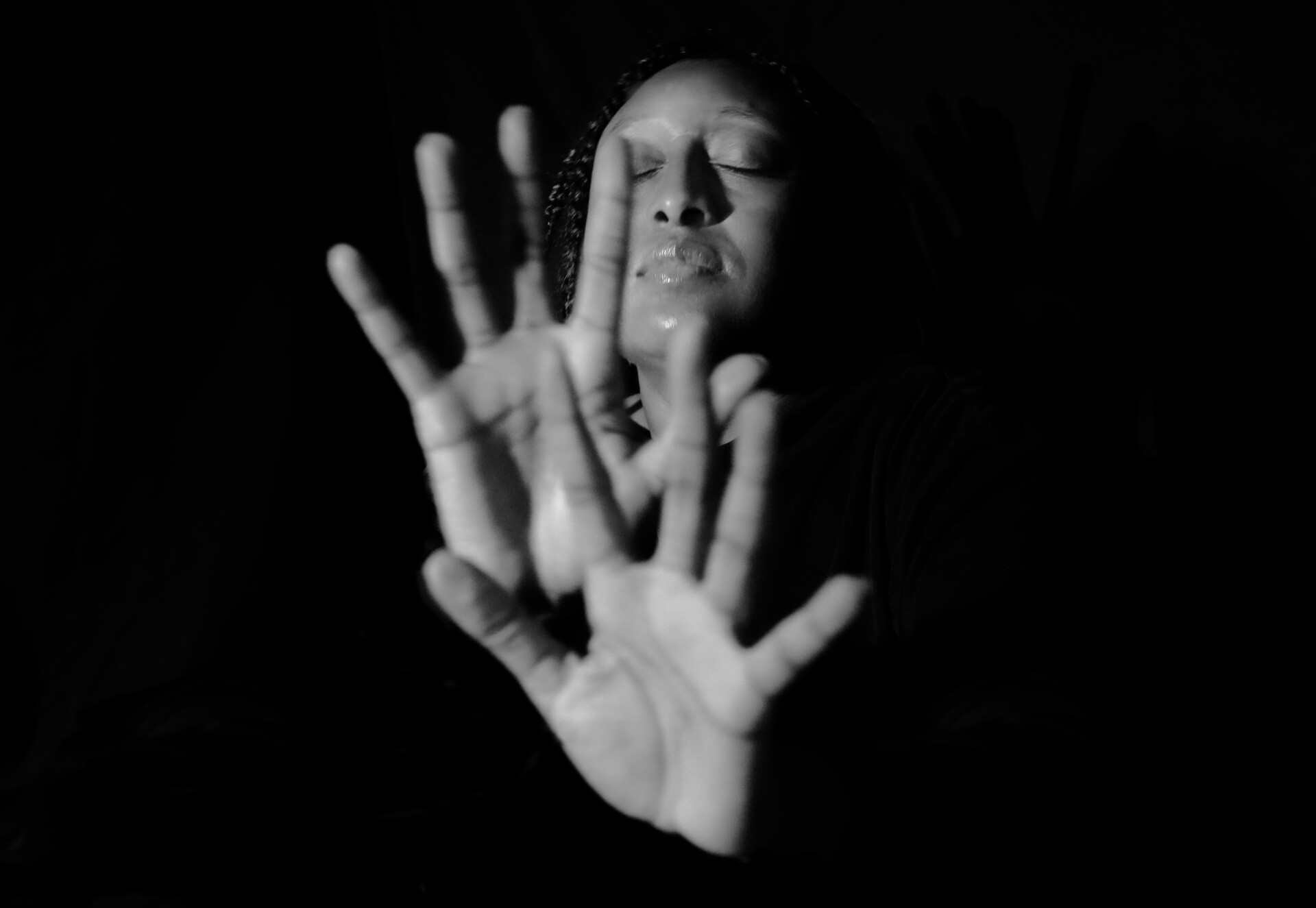 Black and white photograph of a woman with her eyes closed and her hands out in front of her enclosed by a black background.