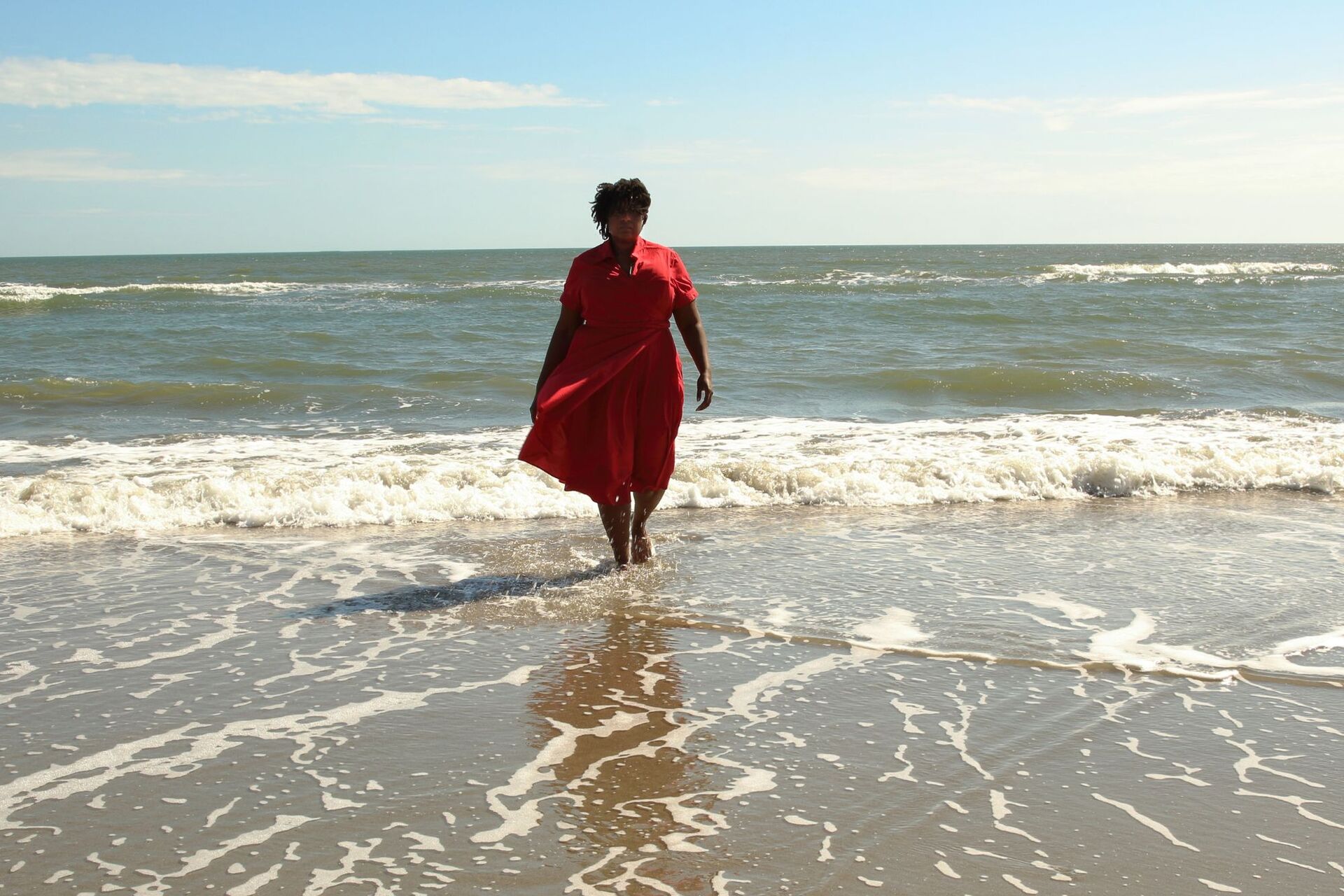 Color photograph of a woman in a red dress walking in the waves on a beach.