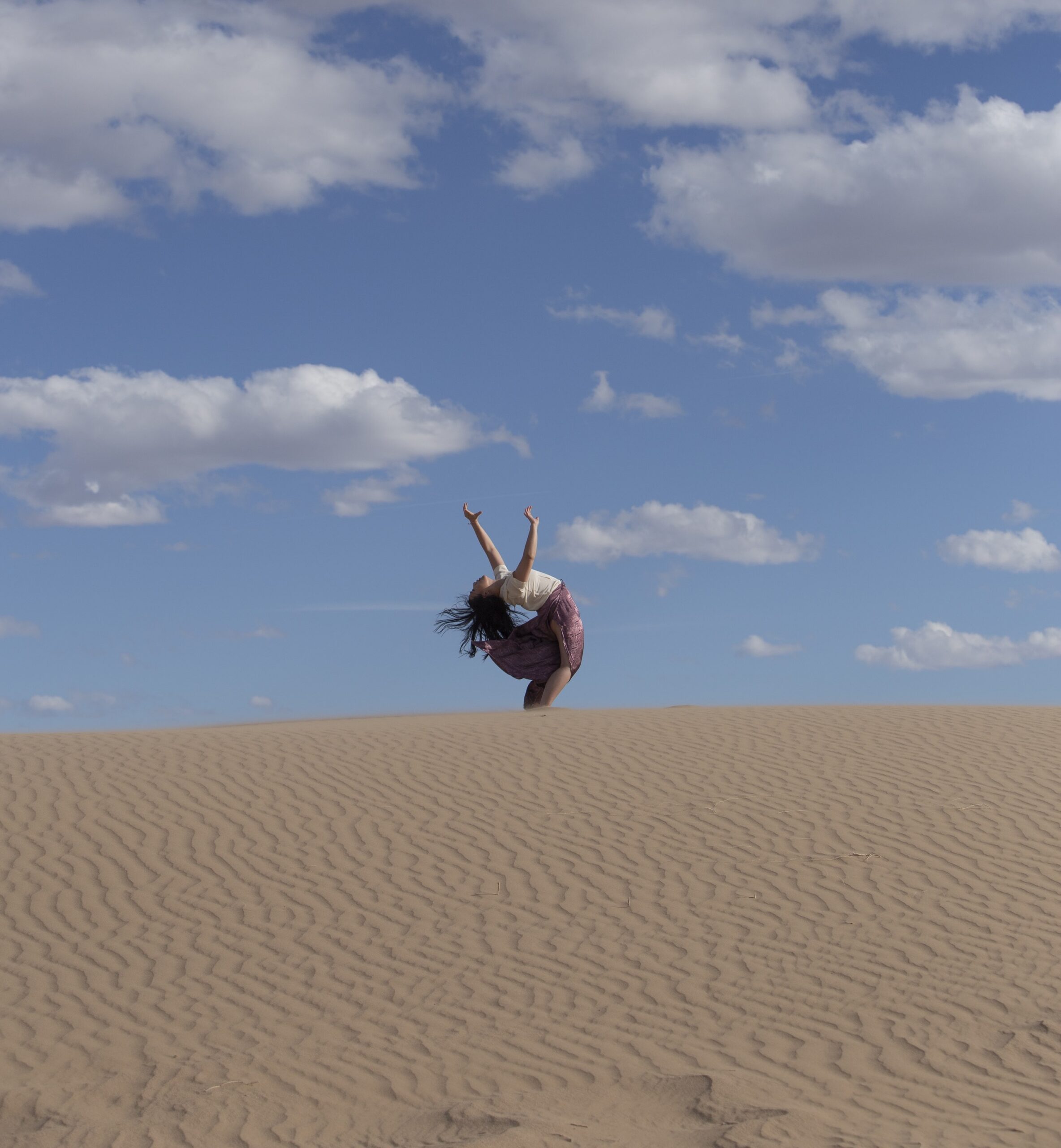 Photograph of a woman on a sand dune bending backwards and reaching her hands towards the sky.