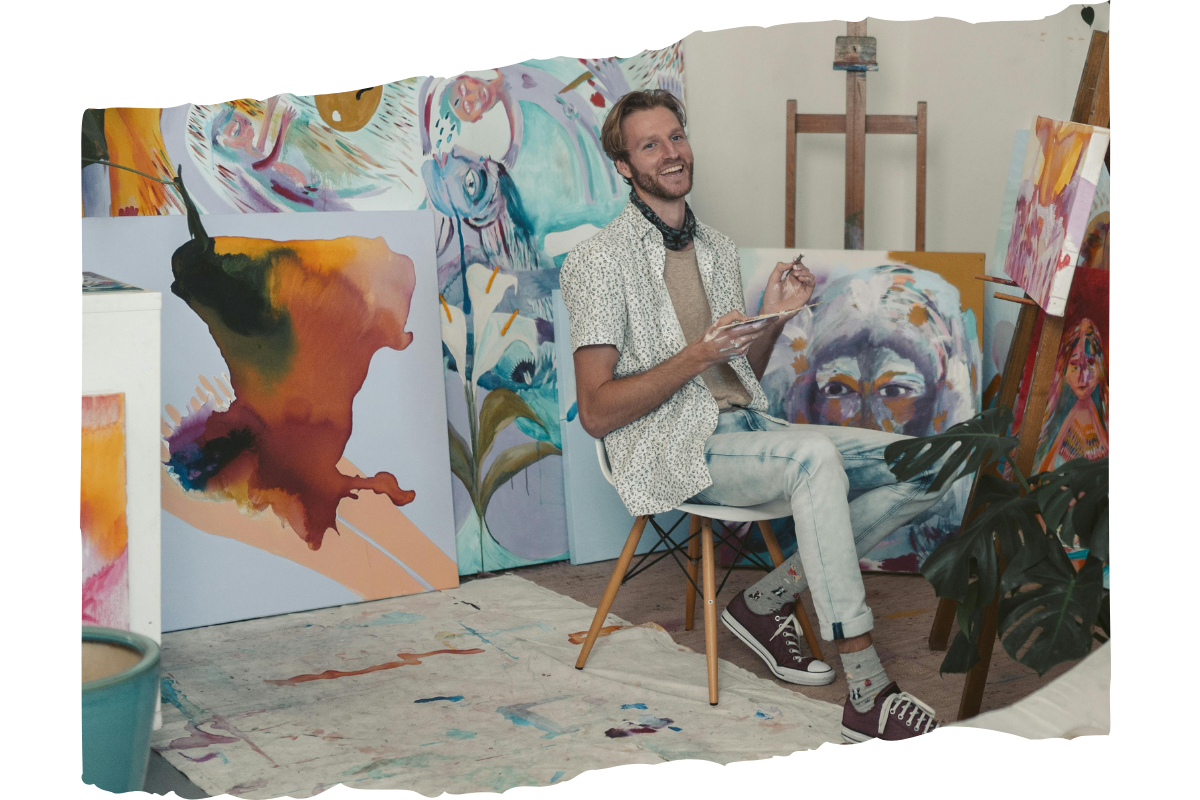 Male artist sitting on a chair holding a paintbrush and smiling while surrounded by paintings in a studio.